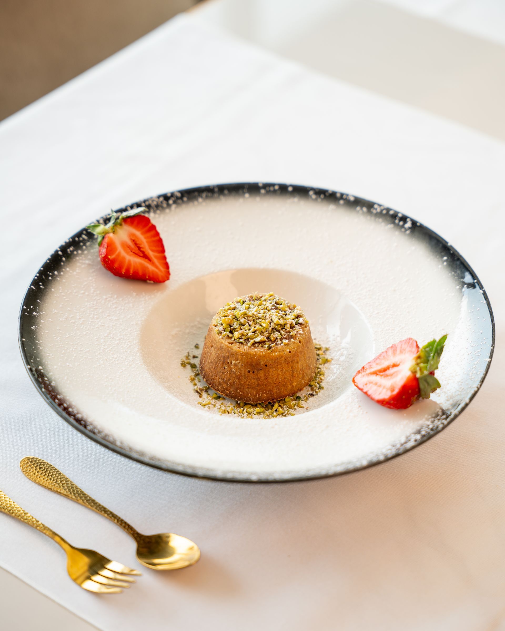 Dessert with pistachio and strawberries on a white plate, with golden fork and spoon.