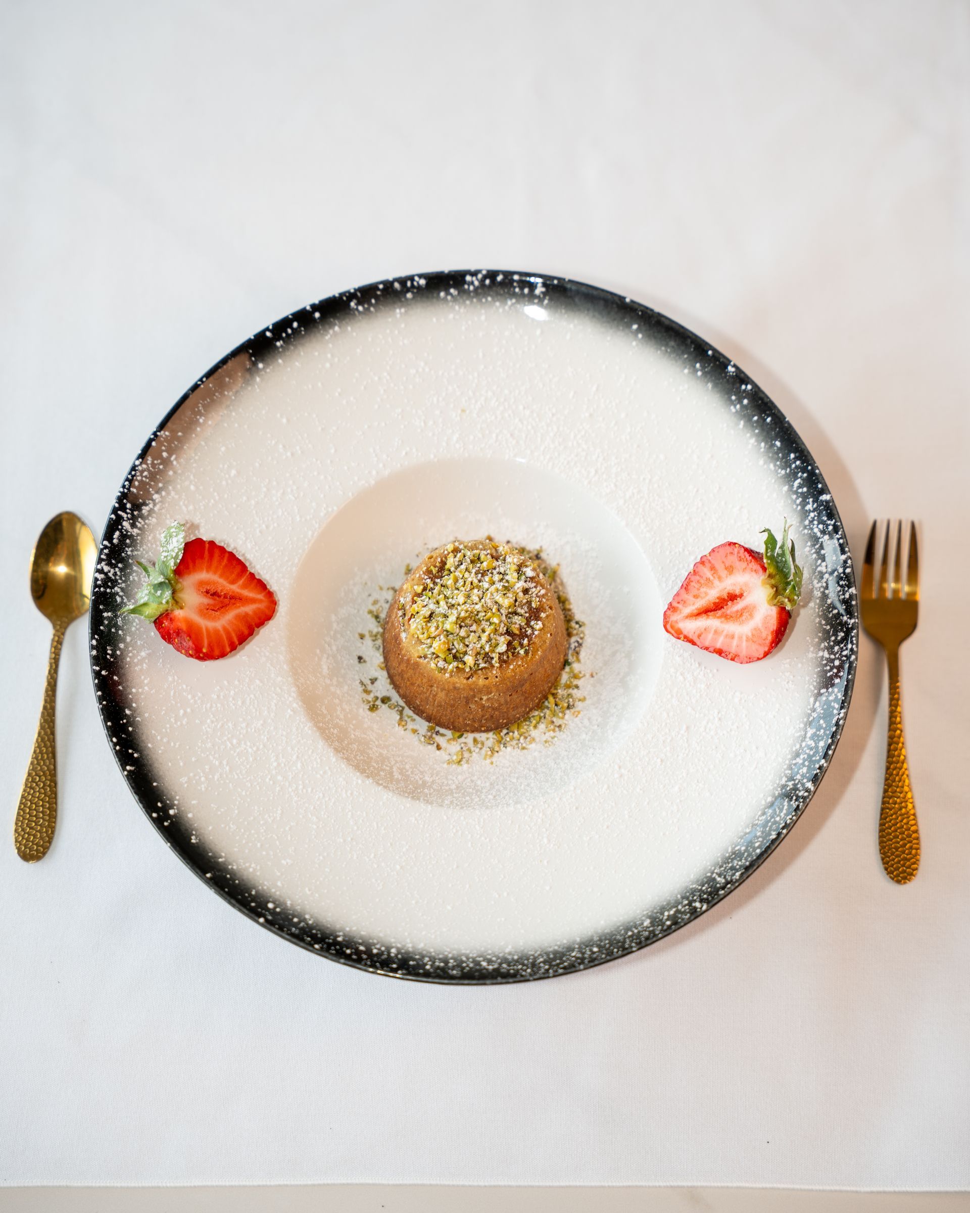Dessert on a plate with gold utensils, two strawberry halves, and sprinkled pistachios.