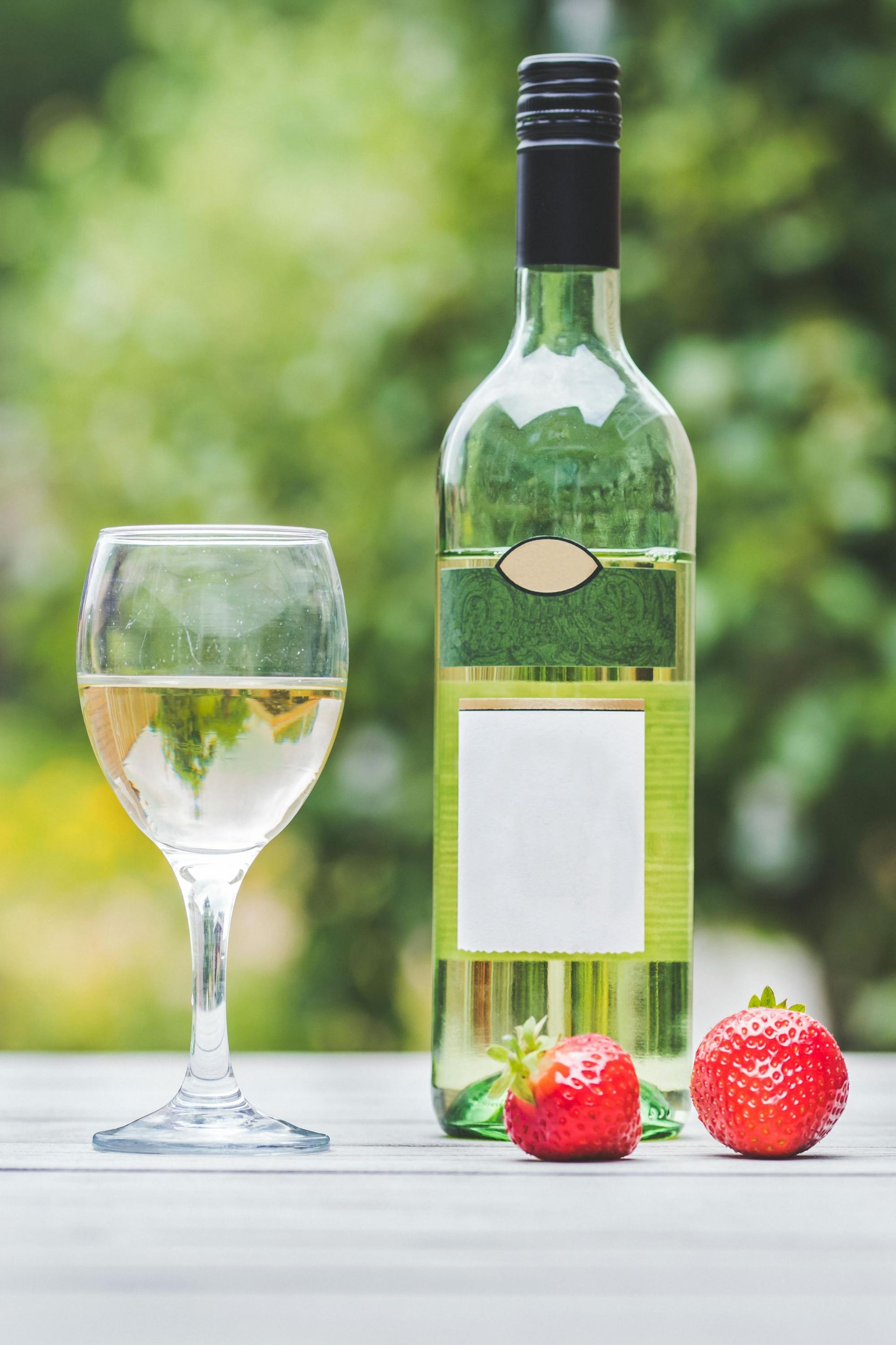 White wine bottle and glass with strawberries on a wooden table, green background.