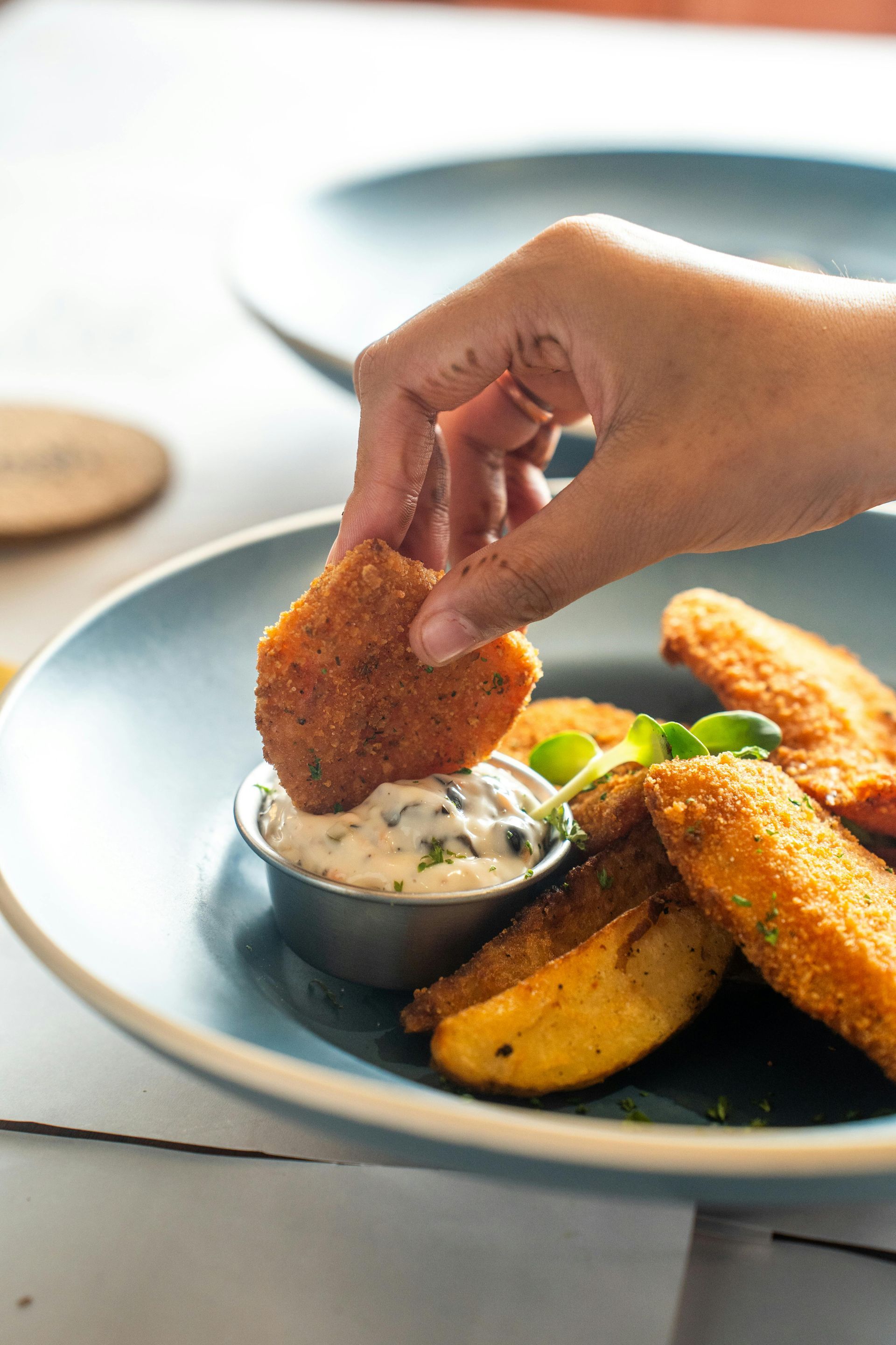 Hand dipping breaded food into white sauce, served with potato wedges on a blue plate.