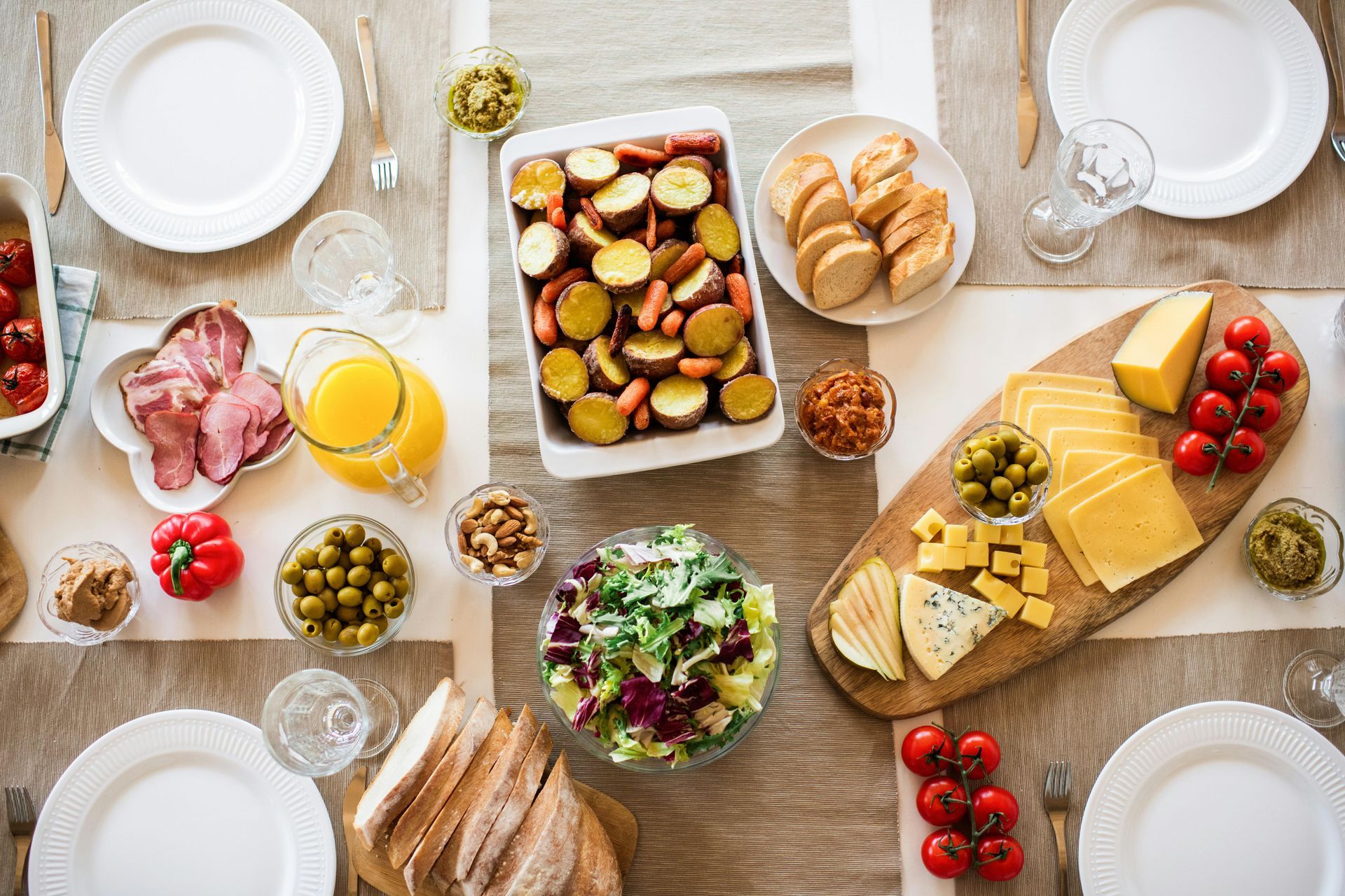 Table set for a meal with ham, potatoes, bread, cheese, salad, olives, tomatoes, and juice.