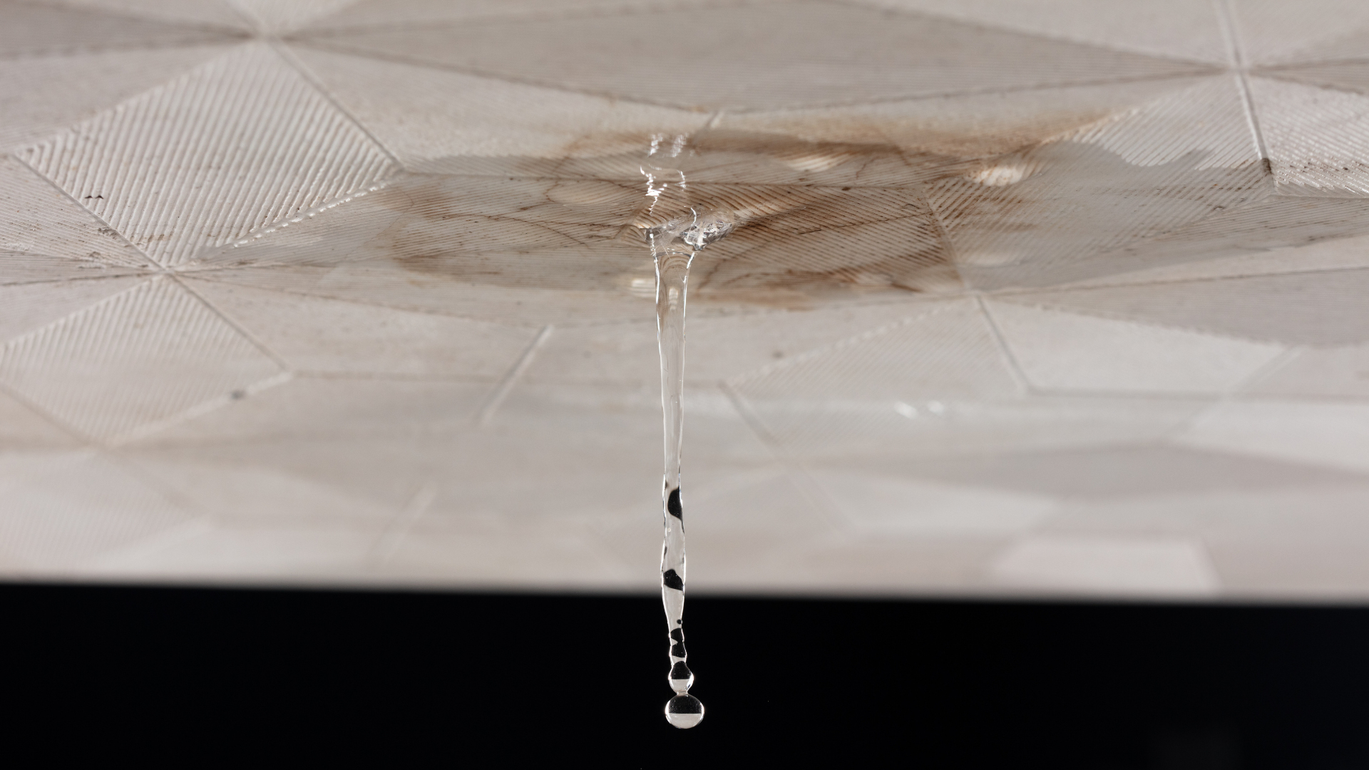 Water drips from a brown, water-damaged spot on a white, patterned drop ceiling panel.