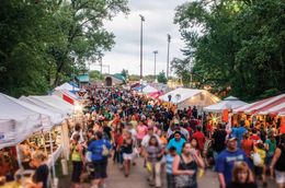 Crowd walking through a fair with white tents, trees, and cloudy sky. People are shopping and gathering.
