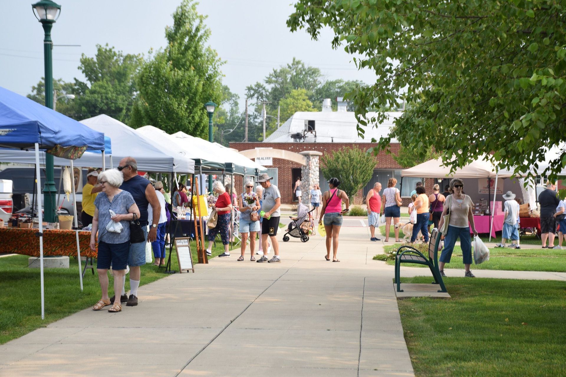 Outdoor farmers market with people browsing stalls under tents on a sunny day.