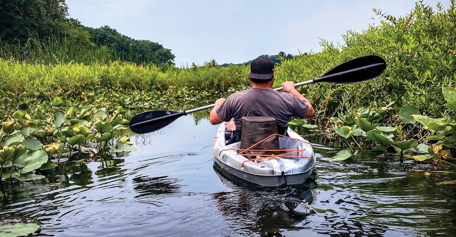 Person kayaking in a narrow waterway, surrounded by lush green foliage and water plants.