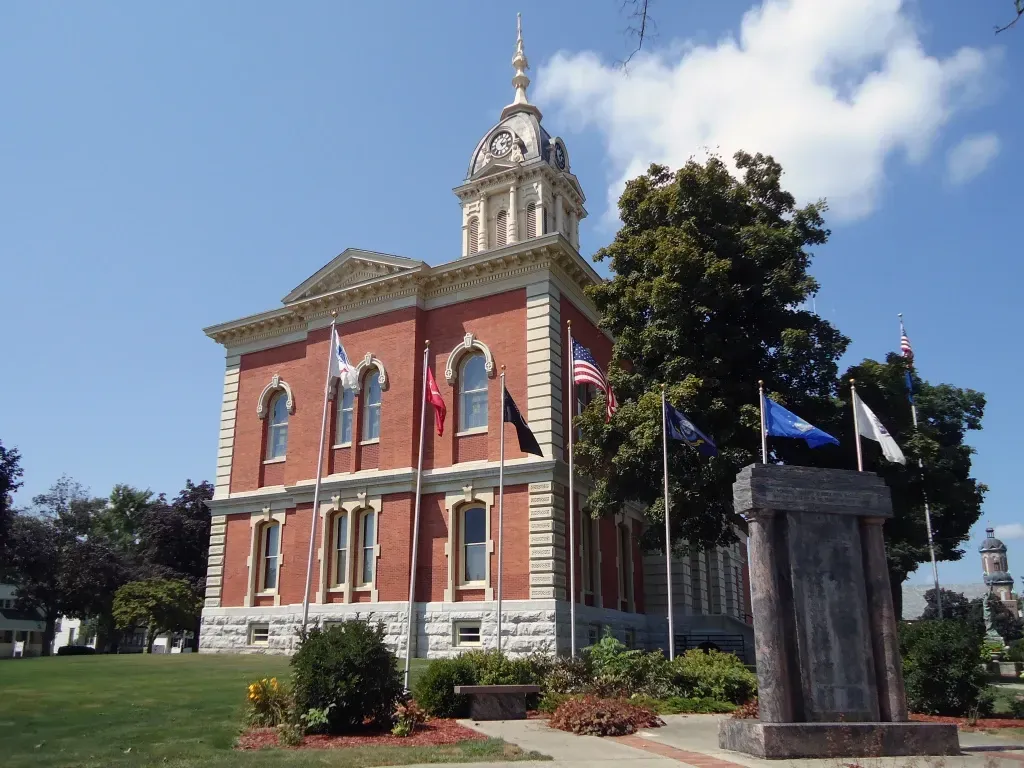 Brick courthouse with a white clock tower and multiple flags on a sunny day.