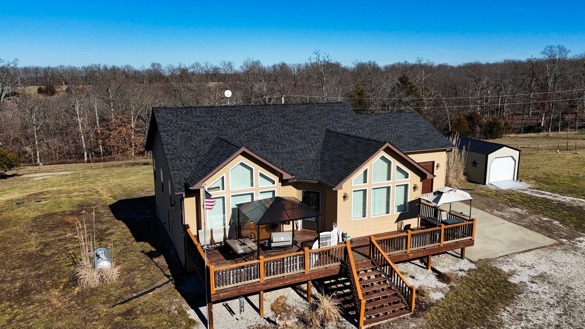 Tan house with a dark roof and a wooden deck, set against a backdrop of bare trees under a clear blue sky.