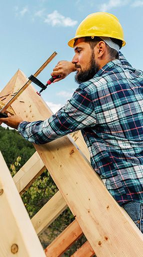 Construction worker in yellow hard hat using a clamp to assemble wooden roof beams.
