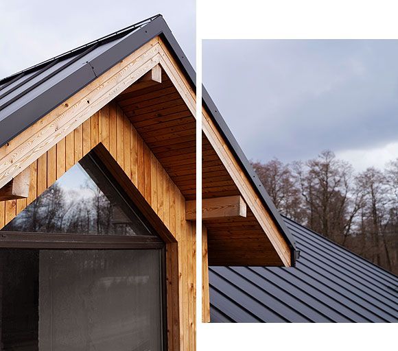 Wood-clad building with black metal roof. Triangular window and a gray sky with trees.