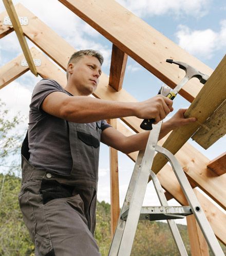 Carpenter on ladder hammering wood on a roof frame outdoors.