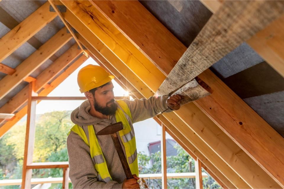 Carpenter in yellow hard hat hammers a board into a roof frame.