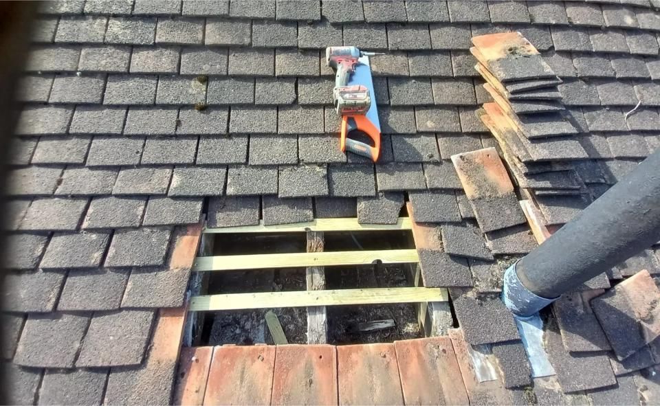 Roof with a hole being repaired, shingles removed. Orange saw, wooden beams, black pipe visible.
