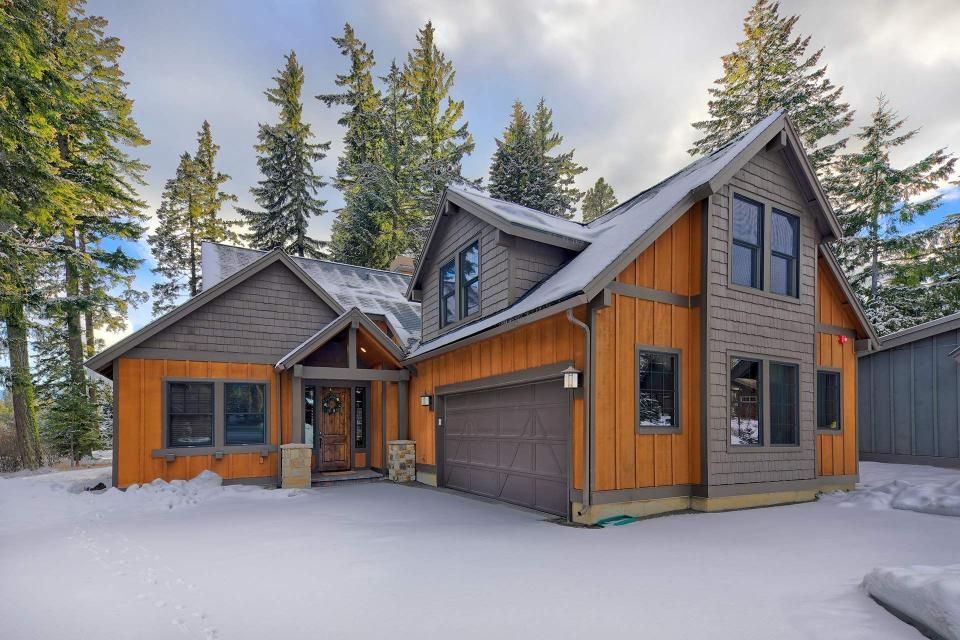 Snow-covered cabin with orange siding, brown trim, and a two-car garage, nestled amongst pine trees.