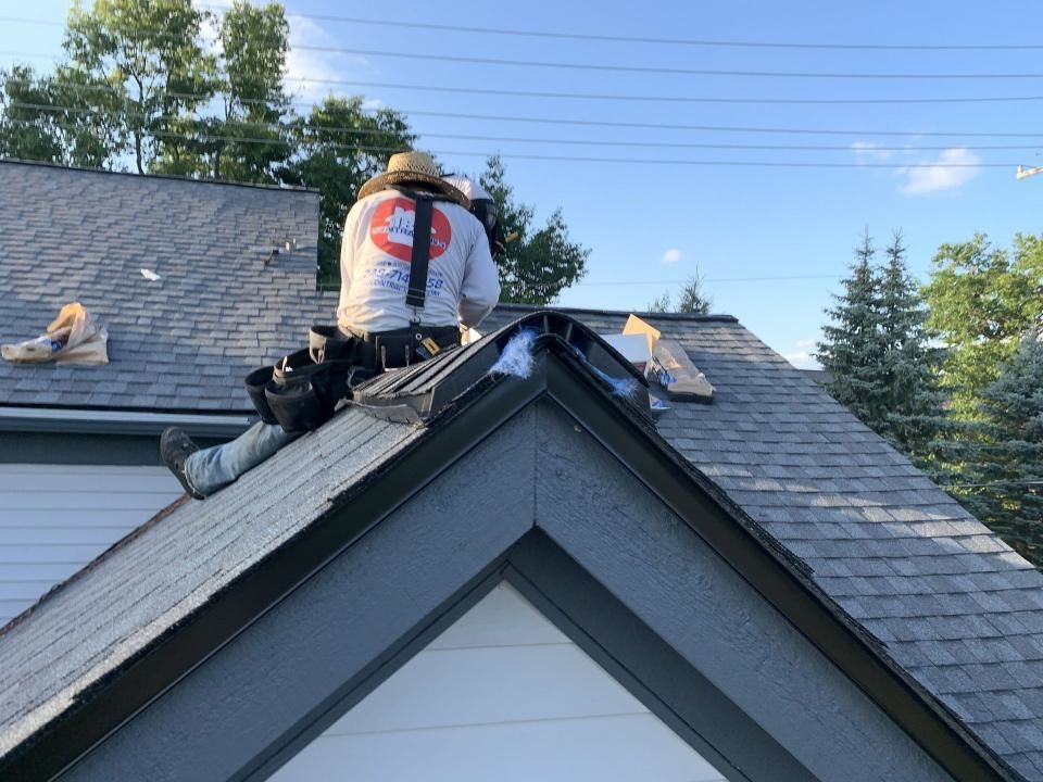 Roofer in safety gear on a roof, installing trim. Bags of supplies and trees are in the background.