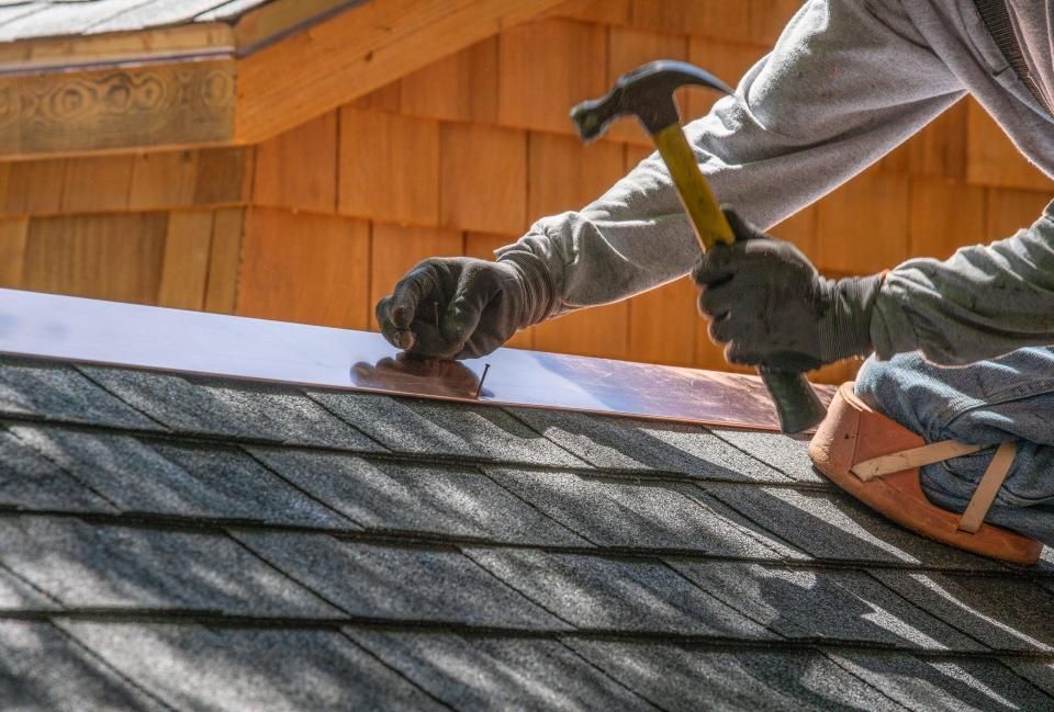 Roofer hammering copper flashing onto a dark shingle roof, wearing gloves and knee pads.
