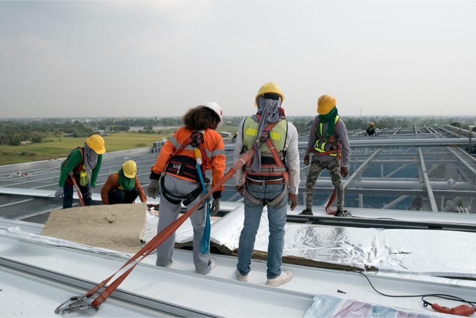 Construction workers wearing safety harnesses working on a roof, sunny day.