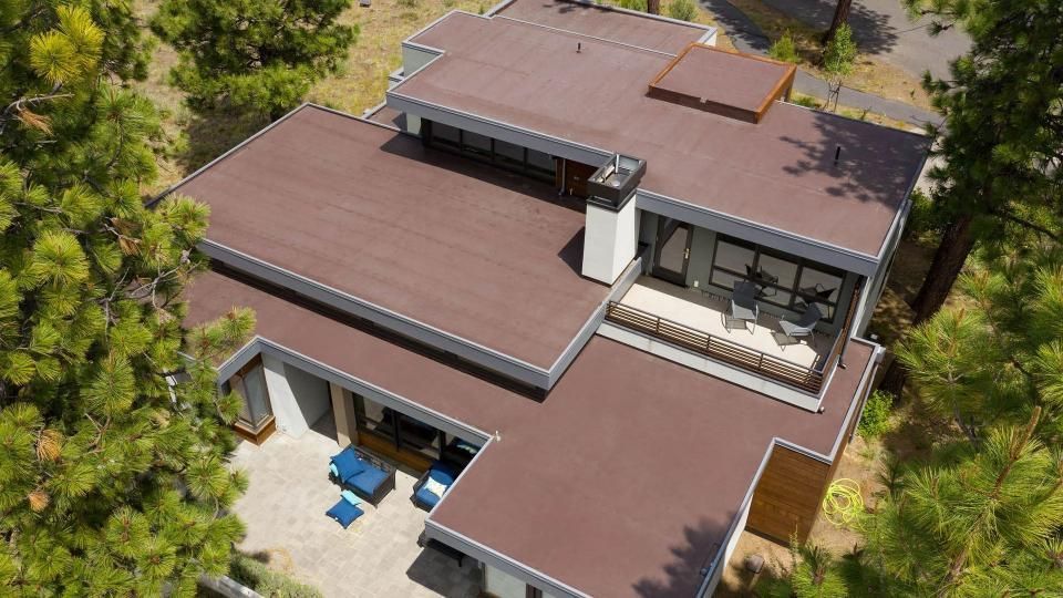 Aerial view of a modern brown-roofed house with outdoor seating and a chimney, surrounded by green trees.