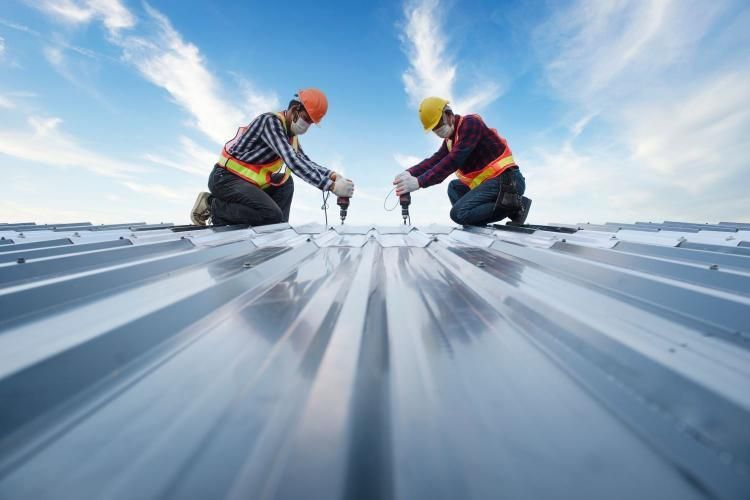 Two roofers in hard hats and safety vests installing a metal roof on a sunny day.