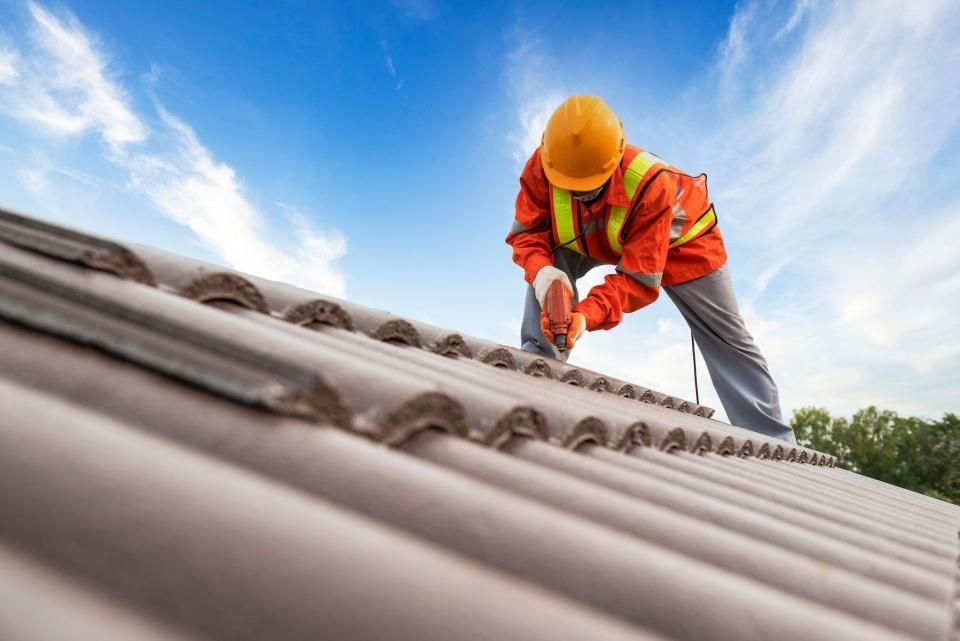 Roofer in orange vest and hard hat using a drill on a tile roof against a blue sky.