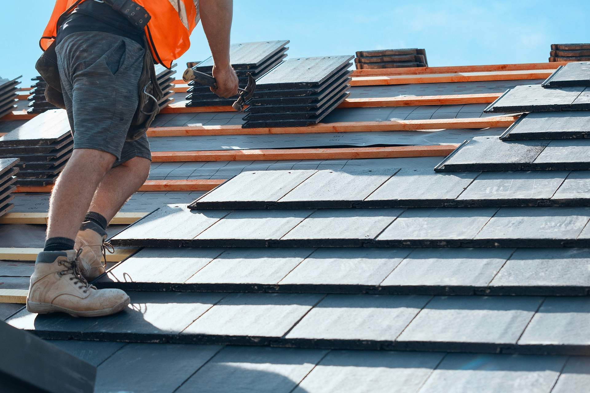 Roofer on a rooftop, laying dark shingles on wooden beams; blue sky in background.