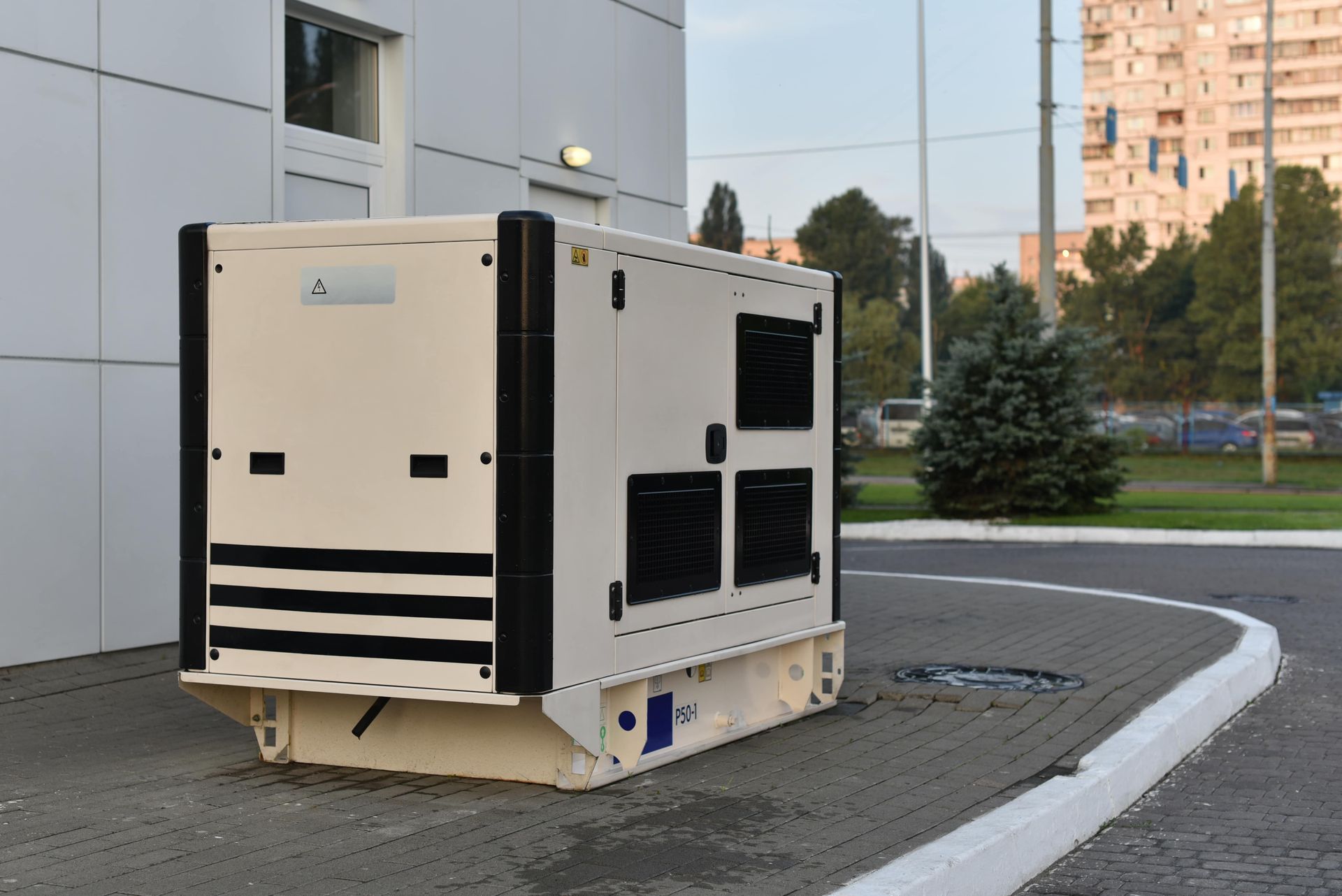 A large, beige generator with black accents sits outside a building on a paved area.