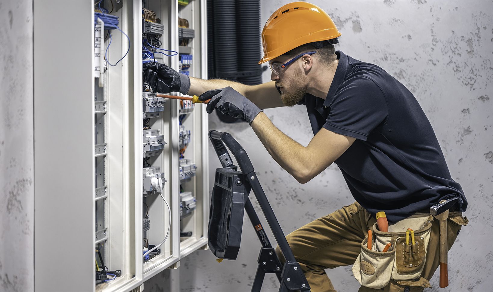 Electrician in hard hat and gloves working on electrical panel.