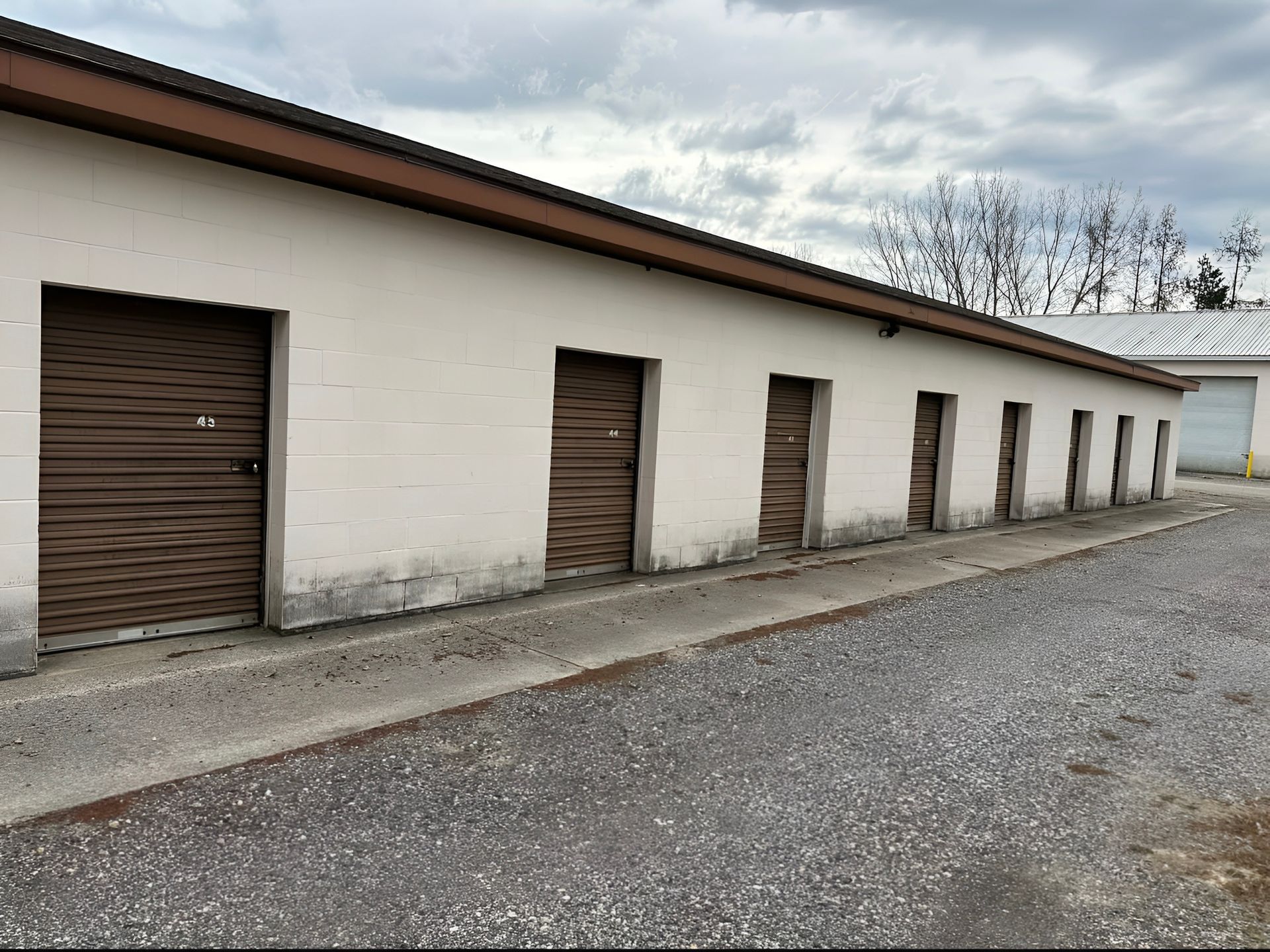 A row of beige storage units with brown doors, along a gravel driveway under a cloudy sky.