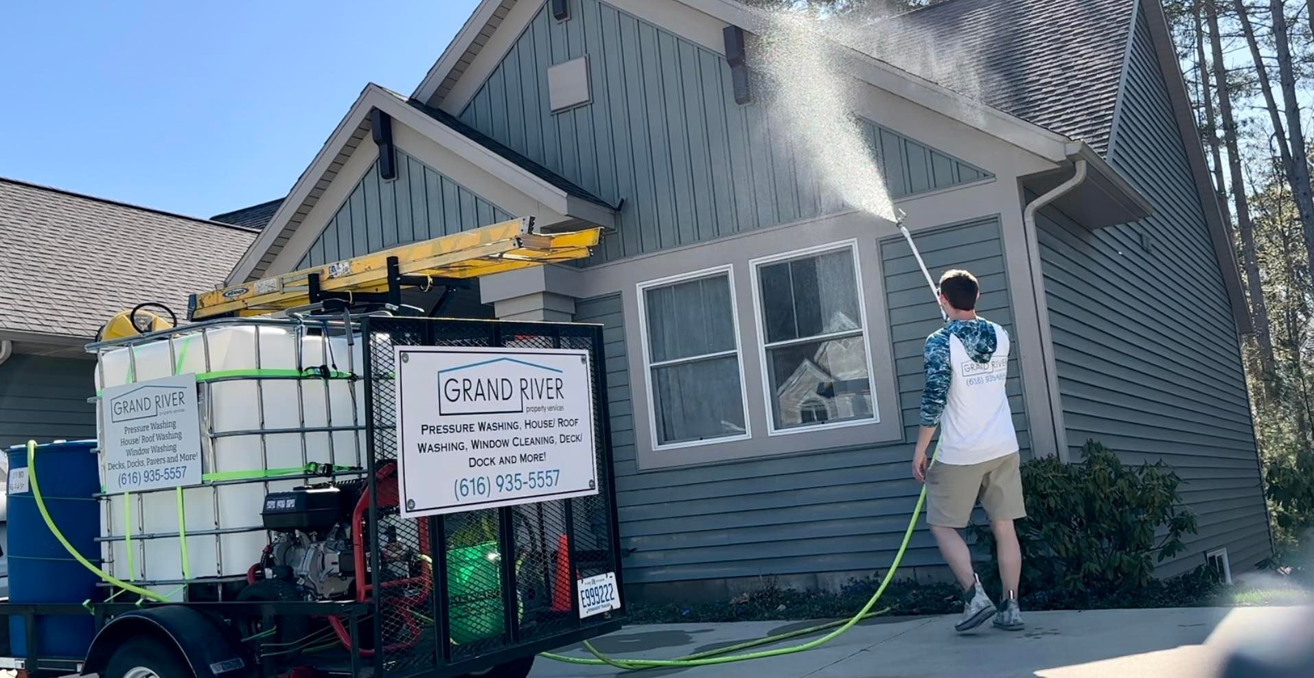 Man power washing the siding of a blue house from a trailer-mounted tank. Sunny day.
