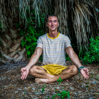 Person meditating cross-legged on the ground before a thatched wall, wearing a striped shirt and yellow shorts.