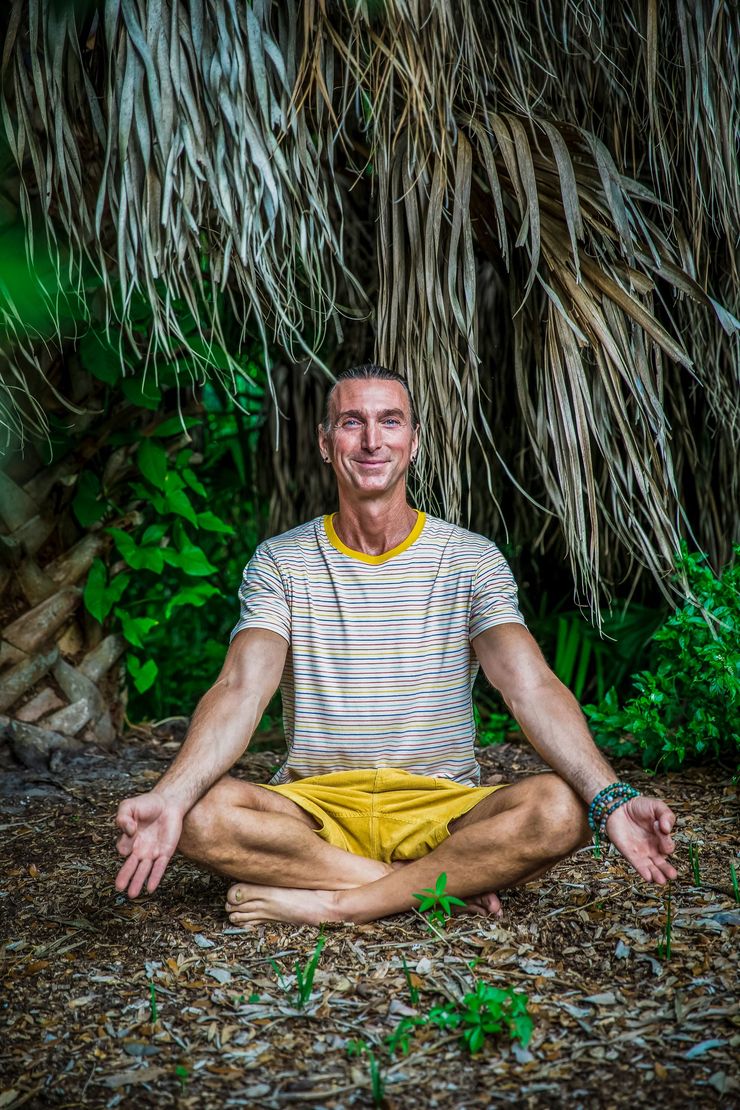 Person meditating cross-legged outdoors against a wall of hanging plants, wearing a striped shirt and yellow shorts