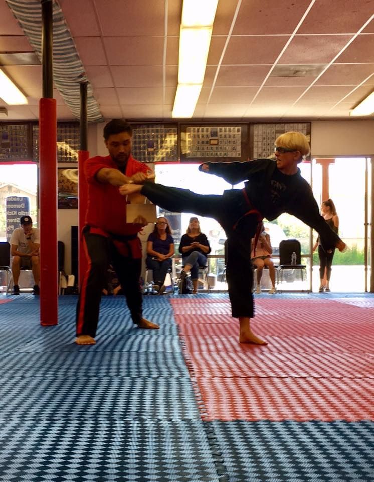 Martial artist kicking a board held by another person in a dojo. Red and blue floor, onlookers.
