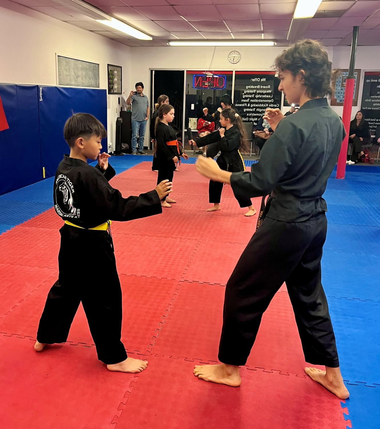 Martial arts students in black uniforms practice stances and techniques on a red and blue mat in a gym studio.