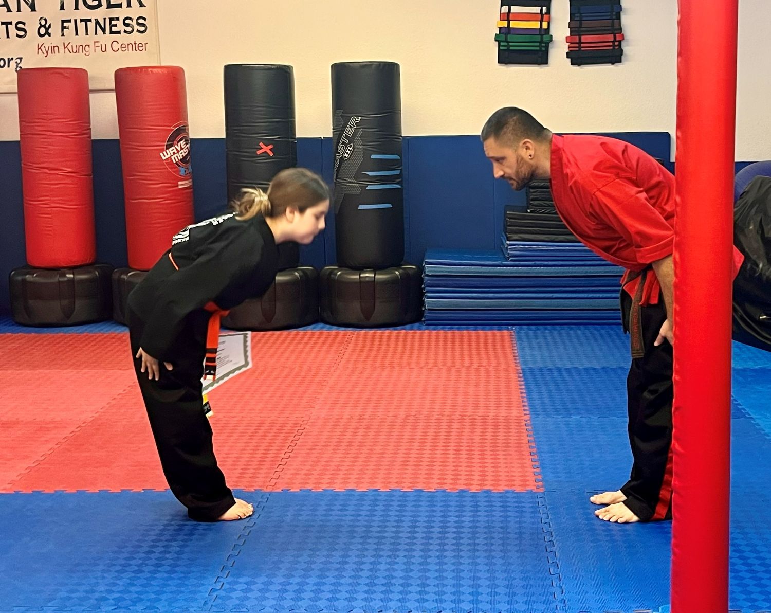 Two martial arts students in uniforms bow to each other on red and blue floor mats in a training facility.