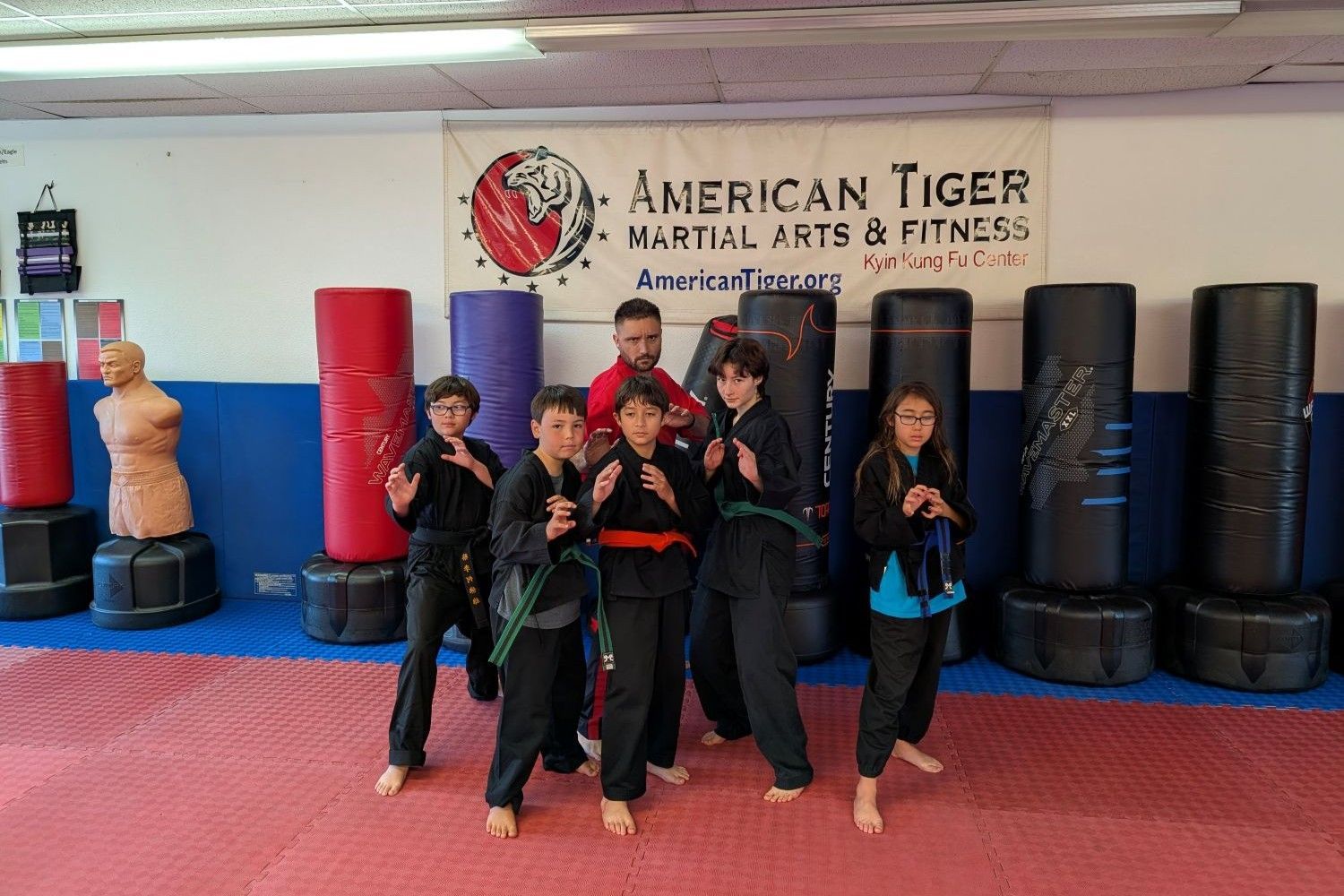 Children in black martial arts uniforms posing in a dojo with punching bags and a sign reading American Tigers Martial Arts & Fitness