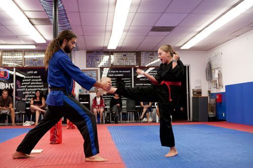 Martial arts training: Instructor in blue uniform, student in black kicking, indoors on mat.