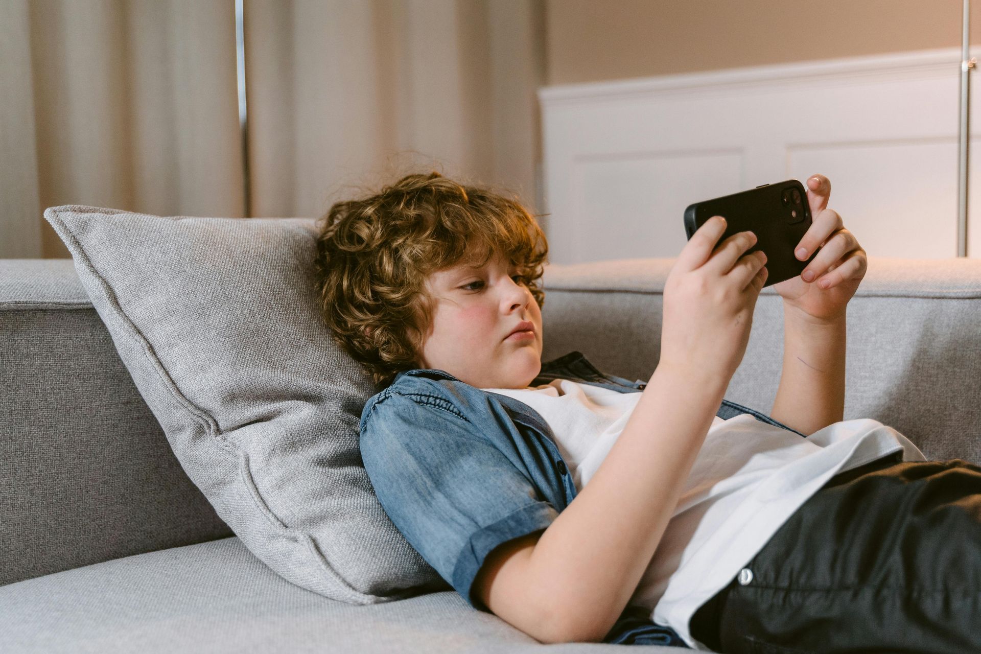 Boy reclining on couch, engrossed in a smartphone. Wearing blue denim jacket, white shirt, dark pants. Soft lighting.