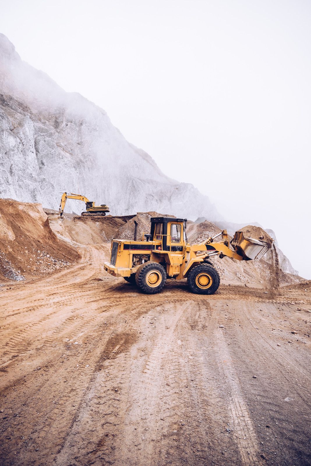 Yellow loader in a quarry, moving dirt. A small excavator works in the background against a cloudy, rocky backdrop.