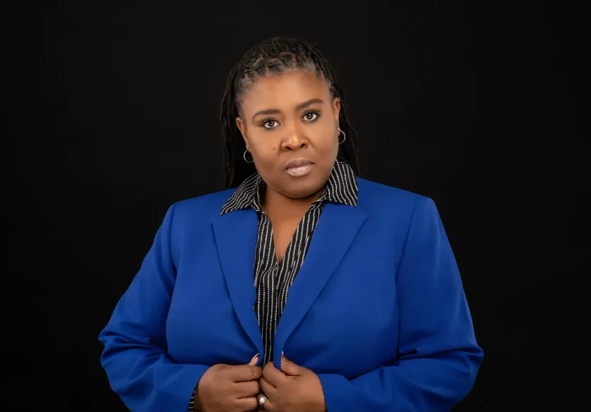 Woman in blue blazer, black and white patterned shirt, stands against a black background, buttoning blazer.