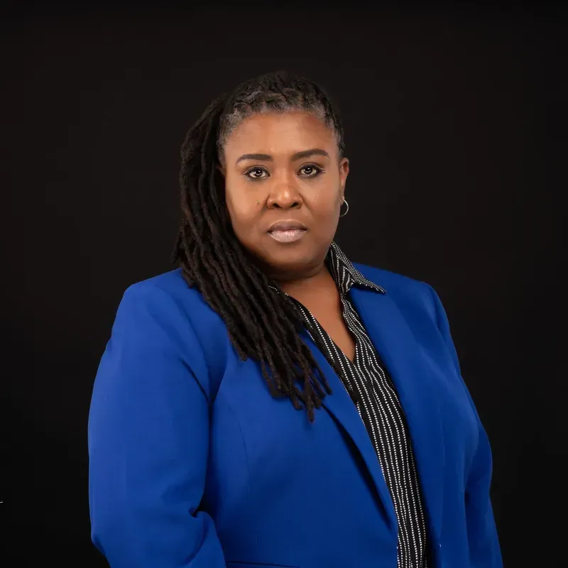 Woman with dark skin, in a blue blazer, against a black backdrop.