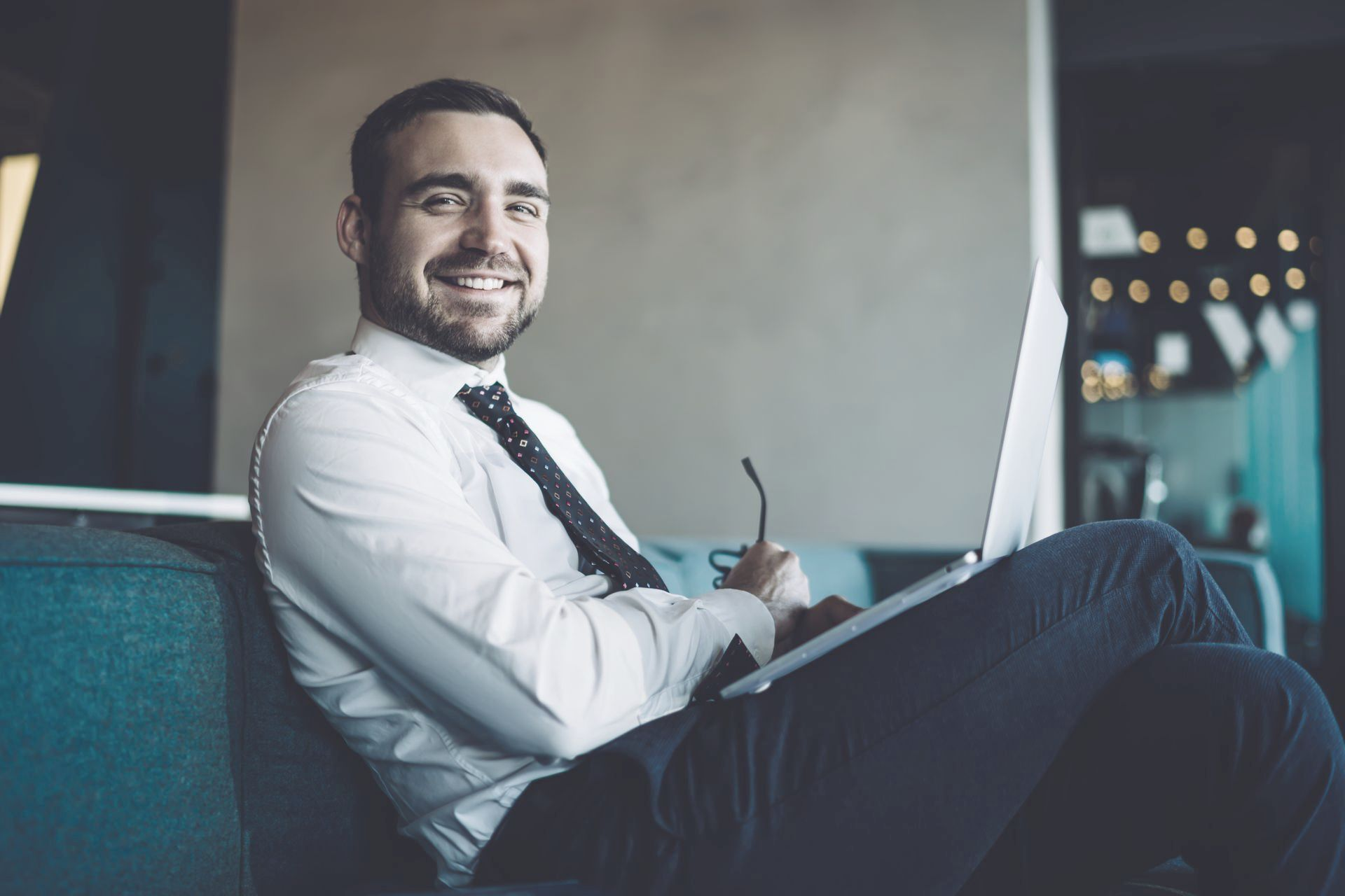 Man in suit sits on couch with laptop, smiling.