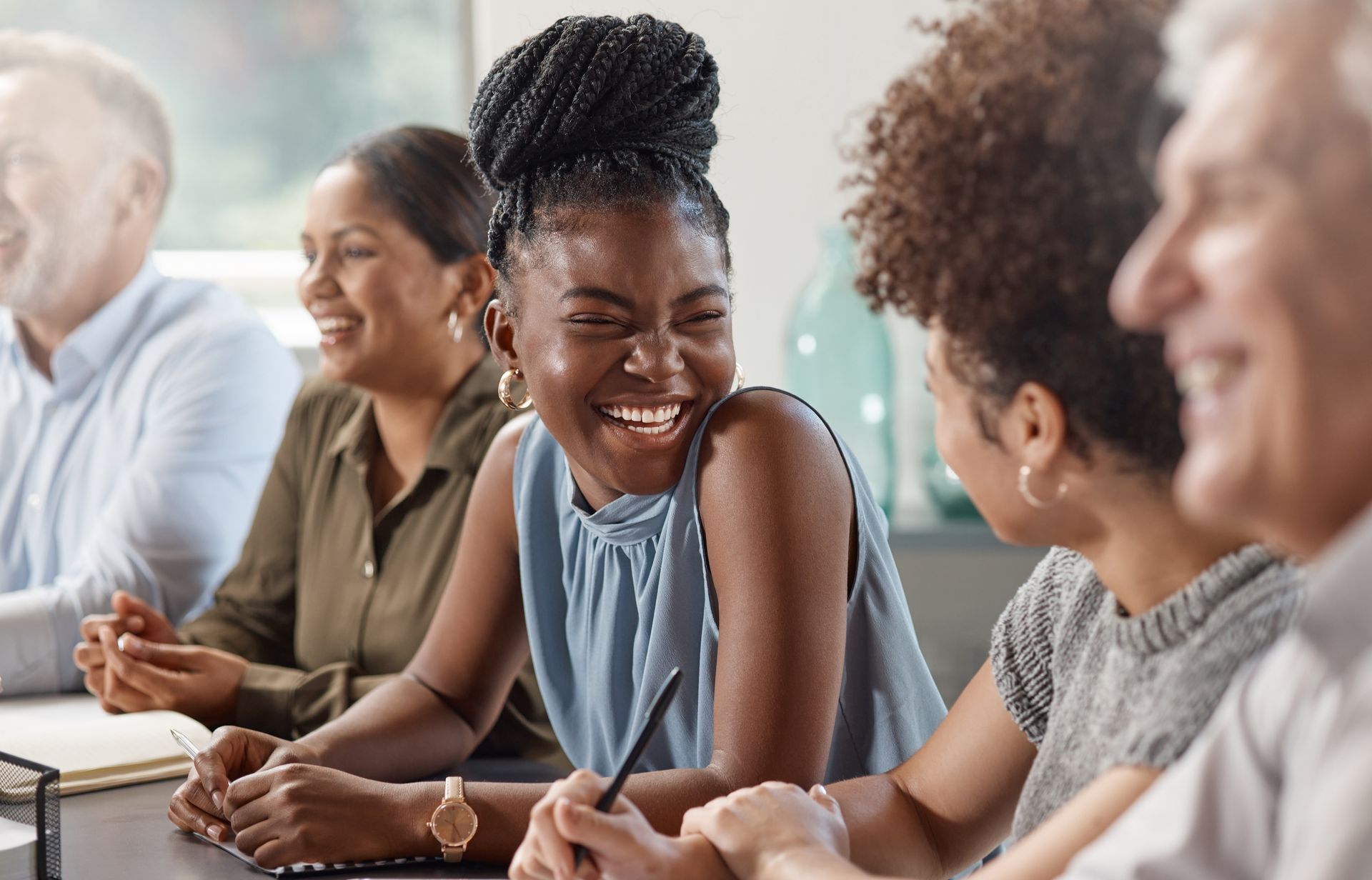 Group of diverse colleagues laughing around a table. Woman in center has large smile.
