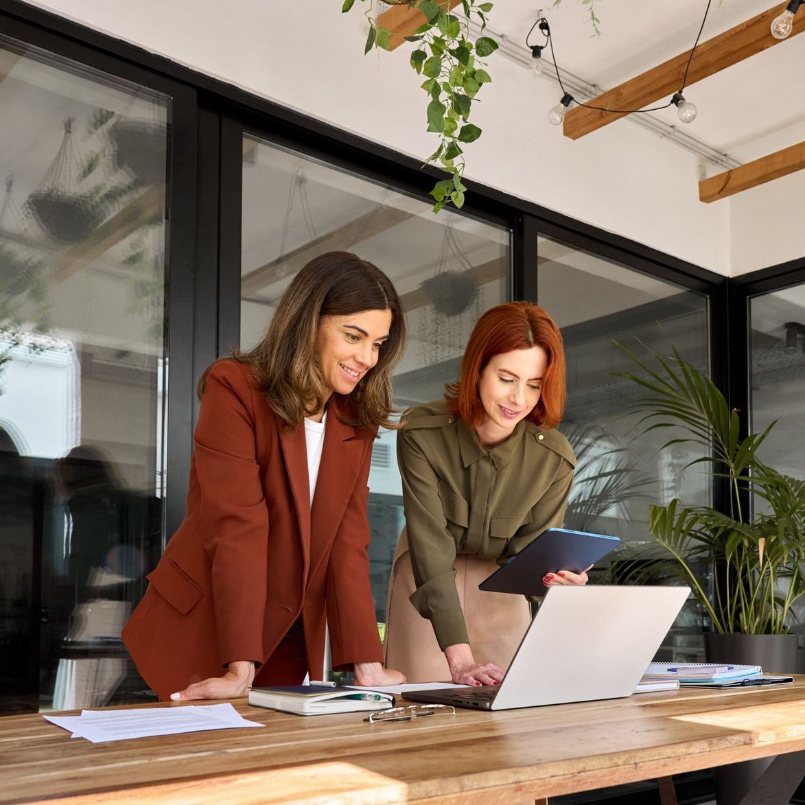 Two women are standing next to each other at a table looking at a laptop.