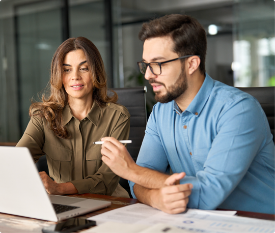 A man and a woman are sitting at a table looking at a laptop computer.