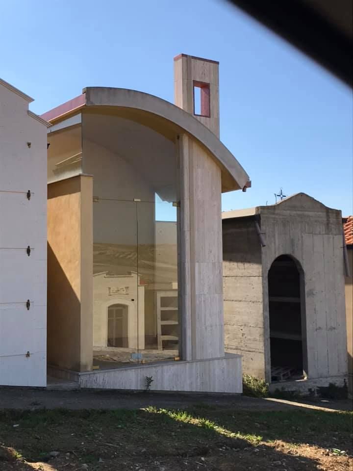 Edificio moderno con ingresso in vetro curvo, cimitero visibile nel riflesso. Giornata di sole, cielo limpido.