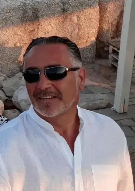 Un uomo che indossa occhiali da sole e una camicia bianca sorride.