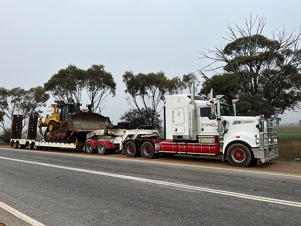 A white semi truck is carrying a bulldozer down a road.