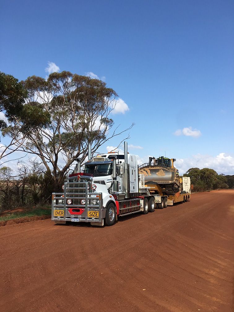 A semi truck is driving down a dirt road with a bulldozer attached to it.