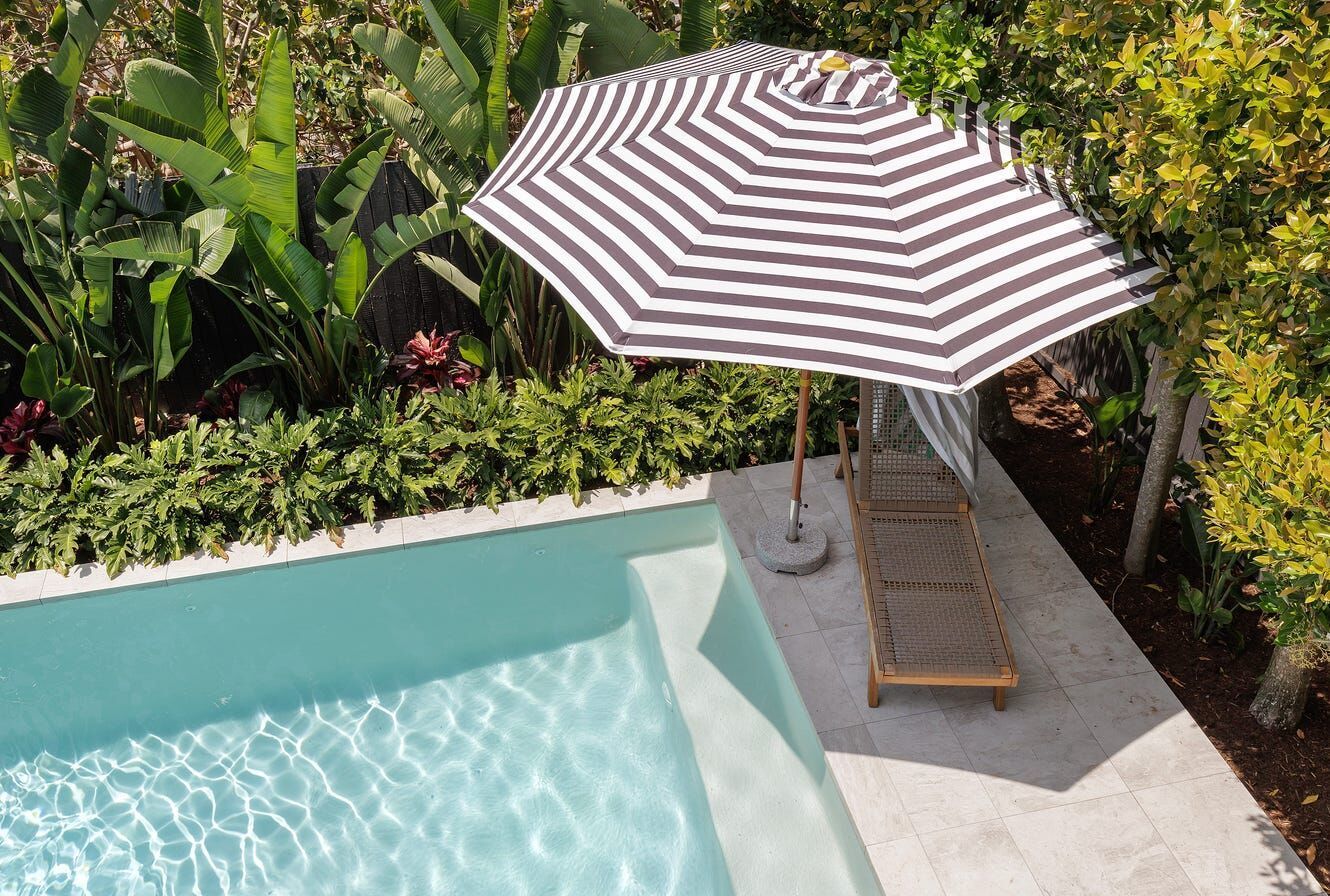 Poolside Scene With Striped Umbrella, Lounge Chair, and Lush Greenery — Mad Pool Interiors and Renovations in Baringa, QLD