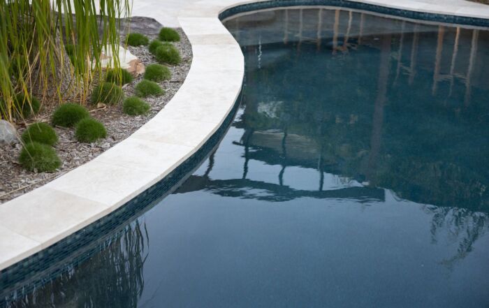 Curved Pool with Dark Blue Water Beside a White Stone Deck and Tropical Landscaping — Mad Pool Interiors and Renovations in Baringa, QLD