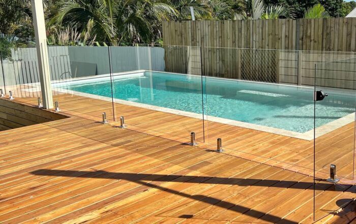 Wooden Deck Beside a Swimming Pool with A Glass Safety Fence and Palm Trees — Mad Pool Interiors and Renovations in Baringa, QLD